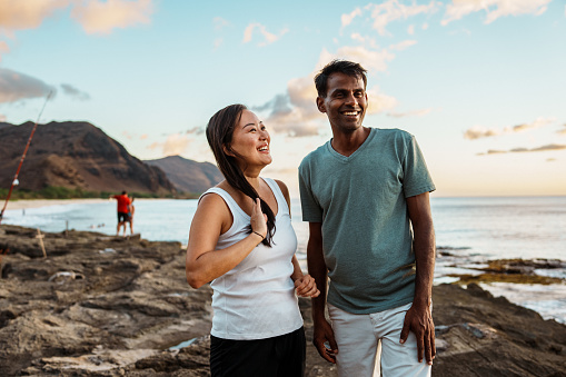 Portrait of an Asian woman and her Indian husband affectionately embracing and smiling directly at the camera while at the beach in Hawaii.