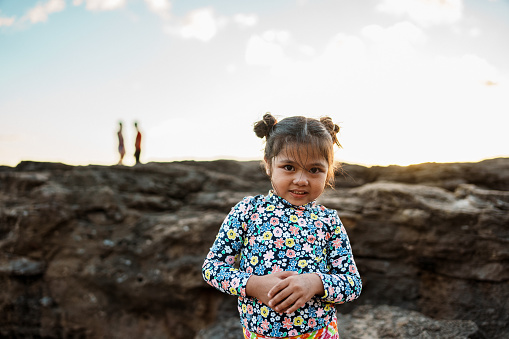 Portrait of an adorable three year old girl of Asian descent standing next to rocks at the beach and smiling directly at the camera.