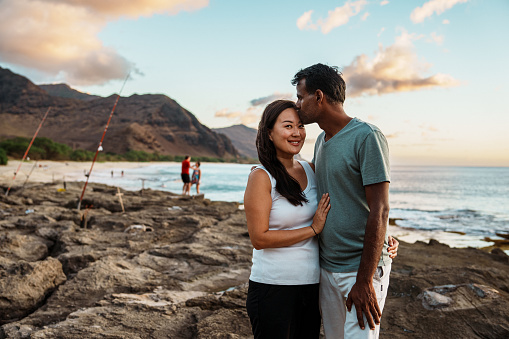 Portrait of an Asian woman and her Indian husband affectionately embracing and smiling directly at the camera while at the beach in Hawaii.