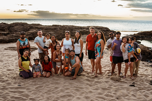 A large multiracial group of adults and children stand on a sandy beach in Hawaii at sunset and smile directly at the camera.