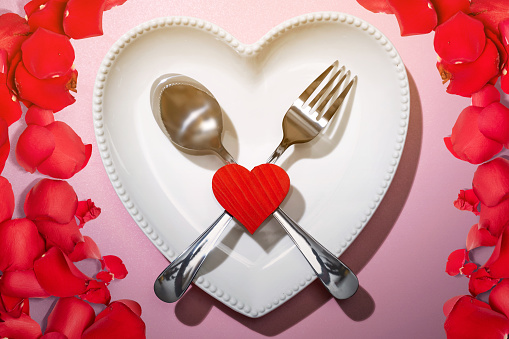 Heart plate with silverware of spoon and fork with a red heart and rose petal over a pink background. Romantic dinner Valentine's Day concept