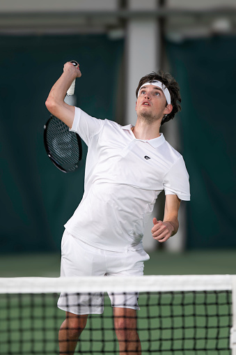 Young man tennis player looking up and hitting ball overhead through net, medium shot front view