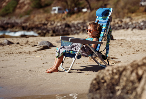 An adorable three year old Eurasian girl wearing a swimsuit and sunglasses relaxes in a folding beach chair on a sandy beach in Hawaii.