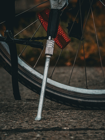 Close-up of a bicycle's rear wheel and kickstand on a textured pavement. The focus is on the metallic kickstand supporting the bike, with a clear view of the wheel, tire, and red reflector. The background is softly blurred, emphasizing the details of the bicycle parts and the asphalt surface