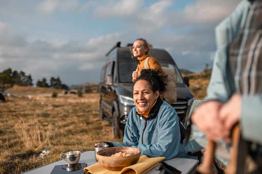 A racially mixed group of hikers meet at a camper, after a hike.
They are sitting in camping chairs, laughing, talking, eating. They greet another group of hikers who come to them, hug, toast with them. They enjoy the sunset and the sun's rays. Sunset, in the mountain grasses on the Gora Nanos plateau in Slovenia. Happy reunion in the mountains