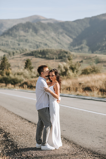 A stylish groom in a white shirt and a cute brunette bride in a white dress are hugging on the side of the road against the background of the forest and mountains. Wedding portrait of newlyweds.