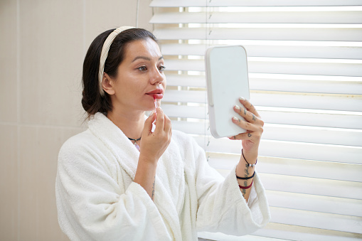 Young woman in white bathrobe looking in mirror and applying red lipstick or lip gloss while standing by window with venetian blinds
