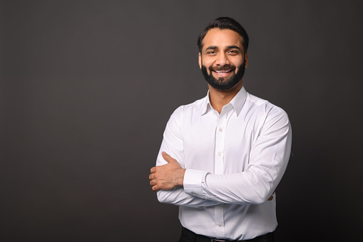 A confident Indian businessman stands with his arms crossed, wearing a crisp white shirt against a black background, exuding professionalism and approachability. Young and successful entrepreneur