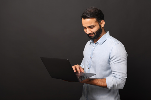 Indian bearded freelancer guy using laptop computer isolated on black background, dark-haired 30s multiracial man in casual shirt typing