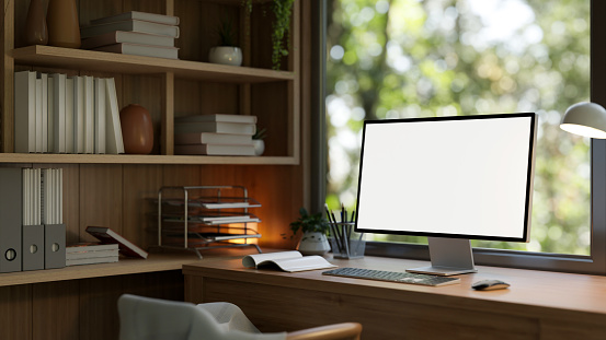 Close-up image of a white-screen computer mockup on a wooden desk against the window in a modern and comfortable home office. 3d render, 3d illustration