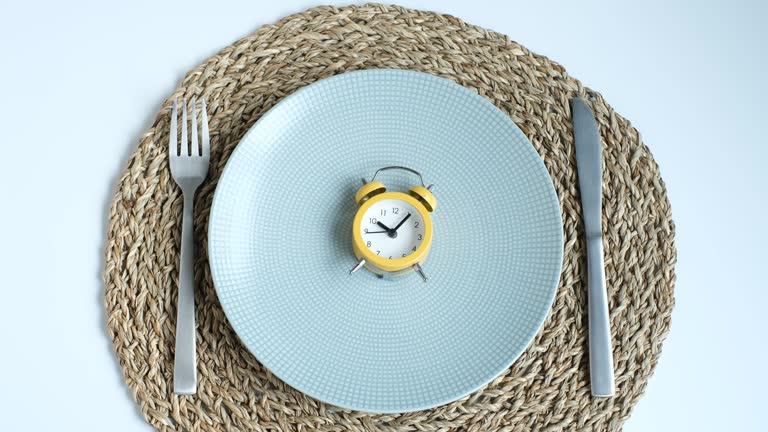 Female hand puts an alarm clock on an empty plate on white background, intermittent fasting concept.