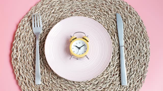 Female hand puts an alarm clock on an empty plate on pink background, intermittent fasting concept.