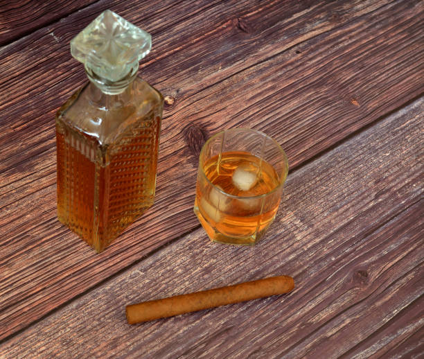 a crystal decanter and a glass of whiskey with ice on a wooden table, next to a cuban cigar. - decanter crystal carafe glass imagens e fotografias de stock