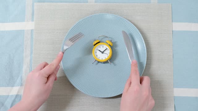 Empty plate with alarm clock and female hand against the background of blue tablecloth, intermittent fasting concept.