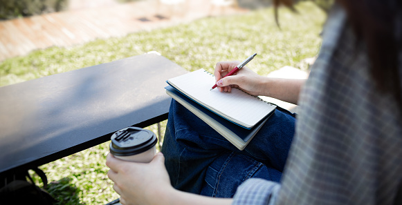 Young asian woman in casual clothes sitting on chair in park taking a note or writing diary in book and other hand holding coffee cup enjoying and relax in her summer holiday
