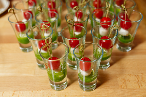 Rows of Caprese salad appetizers with cherry tomatoes, mozzarella cheese, and pesto in clear shot glasses, arranged on a wooden table.