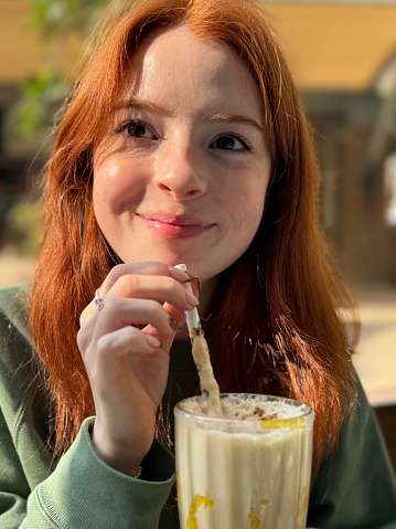 Stock photo showing close-up view of a knickerbocker glass of banana milkshake being drunk by attractive, redheaded woman with a paper drinking straw, in an al fresco cafe restaurant garden setting.