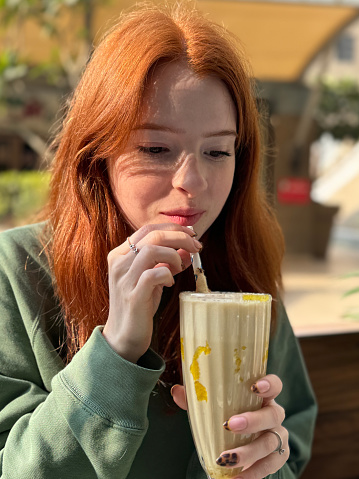 Stock photo showing close-up view of a knickerbocker glass of banana milkshake being drunk by attractive, redheaded woman with a paper drinking straw, in an al fresco cafe restaurant garden setting.