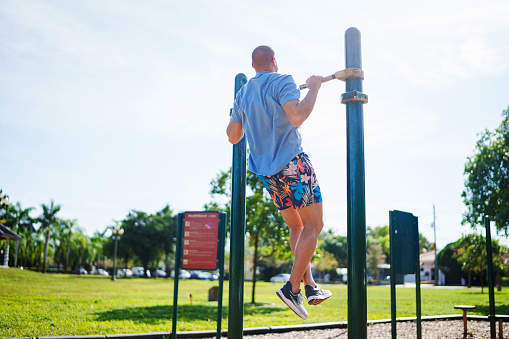 A healthy and fit man of Caucasian descent does a pull-up while exercising in a public park on a beautiful, sunny day in Florida.