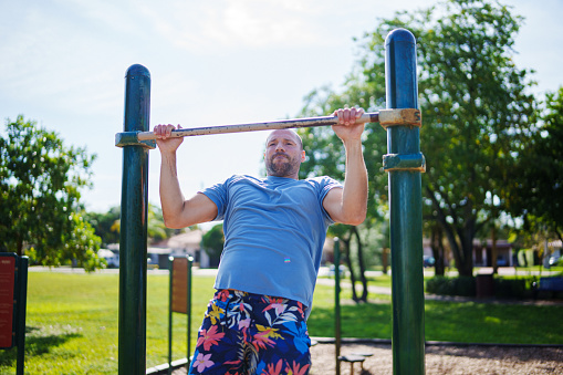 A healthy and fit man of Caucasian descent concentrates while doing a pull-up and exercising in a public park on a beautiful, sunny day in Florida.
