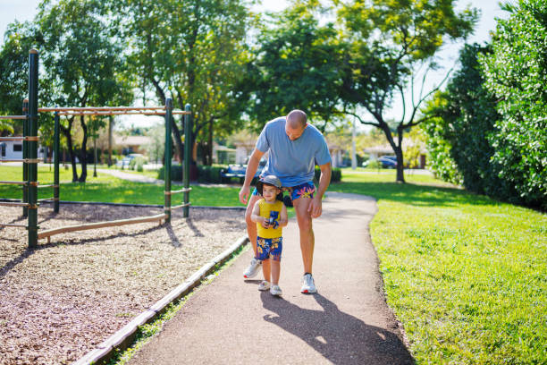 A middle aged Caucasian father walks with his Puerto Rican toddler son on a sidewalk while enjoying a beautiful day in a Florida park together.