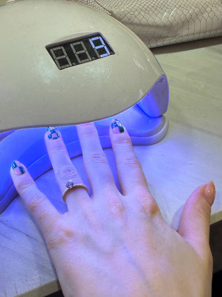 istock Close-up image of unrecognisable woman's left hands displaying nail art and engagement ring, fingers spread under nail curing lamp showing green checked pattern nail varnish manicure design, manicure drying under uv light in nail salon, elevated view 1994309633