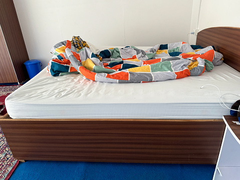 Stock photo showing an empty, unmade double bed with crumpled, white bedding. In the bedroom beside the bed stands a wooden bedside table and wardrobe.