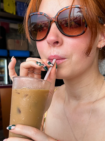 Stock photo showing close-up view of iced coffee in glass being drunk by red haired woman through drinking straw at an al fresco restaurant table.