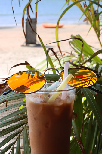 Stock photo showing close-up image of al fresco beach bar restaurant scene with a glass of iced coffee with ice cubes and three drinking straws. Pictured is an iced coffee with ice cubes in drinking glass covered in condensation.