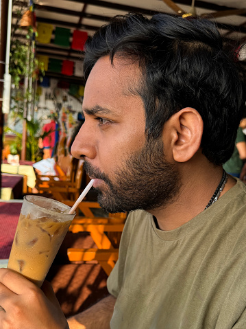 Stock photo showing close-up view of iced coffee in glass being drunk by an Indian man through drinking straw at an al fresco restaurant table.