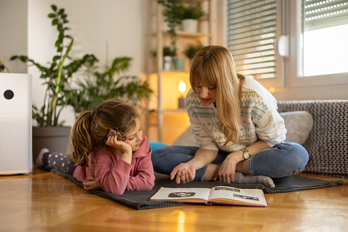 Devoted mother reading a book to her little daughter while sitting on floor at home