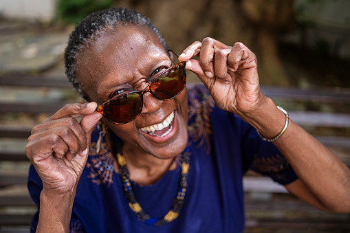 Portrait of a stylish senior woman looking over the rim of her sunglasses and laughing while sitting outside on a park bench in summer