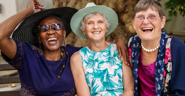 Smiling senior women sitting together outside on a park bench Portrait of three diverse senior female friends laughing while sitting together on a bench in a park in summer 70 79 years stock pictures, royalty-free photos & images