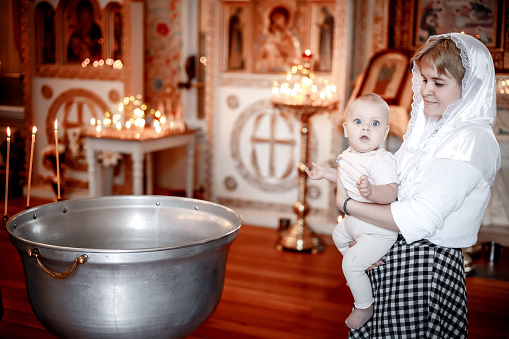 the sacrament of the baptism of a child, a mother with a small child praying in an Orthodox Christian church or temple, believers at the font and icons.