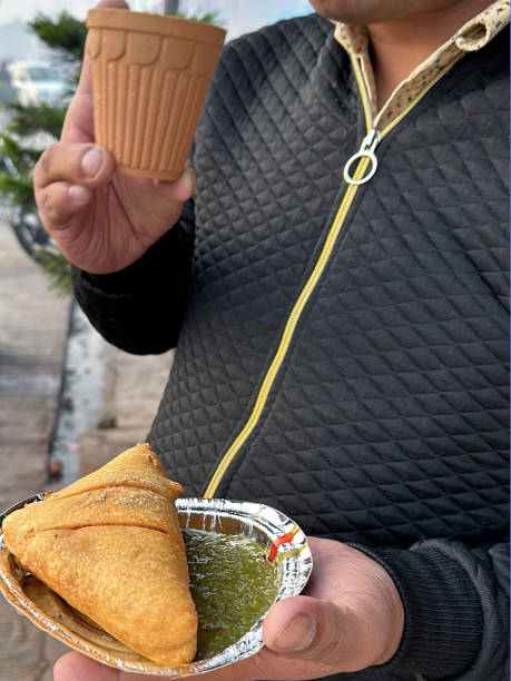istock Full frame image of unrecognisable man holding disposable foil plate of samosa stuffed with spiced potato, peas and meat served with mint coriander dip and clay beaker of masala chai tea from street food market stall, focus on foreground 1994454553