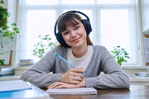 Web camera view of teenage student girl in headphones looking at camera sitting at desk with textbooks notebooks. Online lesson video chat call conference. Technology education training e-learning