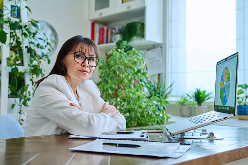 Portrait of mature businesswoman working at home on computer laptop. Smiling middle-aged female looking at camera sitting at desk in home office. Remote business work, career, management, marketing