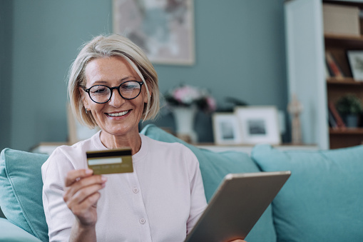 Cheerful woman shopping online at home. Shot of mature woman shopping online with credit card and digital tablet.