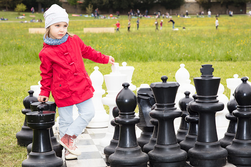 A little girl is playing chess in a public park. She is thinking about her next move, leaning on huge pawns.