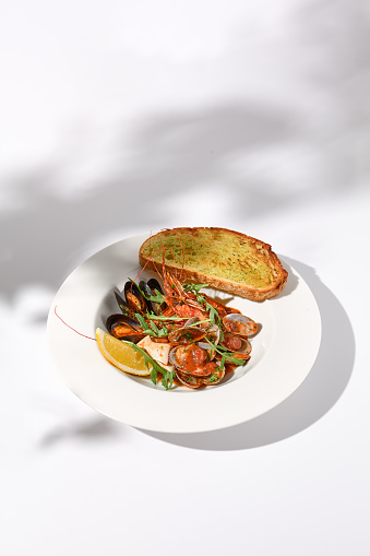 SautÃ©ed shellfish medley with ciabatta toast, side view on white plate with leaf shadow art.