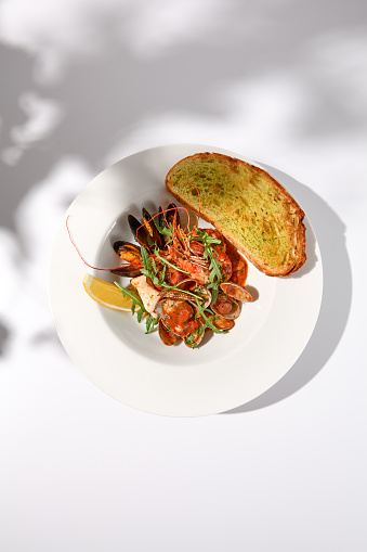 SautÃ©ed shellfish in tomato sauce with garlic ciabatta toast, top view on white with dramatic shadow play.