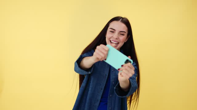 Carefree happy woman wearing casual denim shirt playing video games on modern smartphone in studio. Attractive female with brown hair having fun during online entertainment over yellow background.