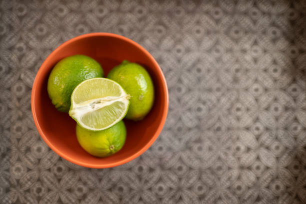 A top view of limes in an orange bowl on a silver-patterned background, empty right-hand side space