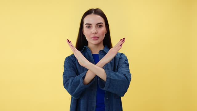 Portrait of young woman with serious facial expression crossing hands for showing stop gesture. Annoyed brunette refusing something with sign of prohibition. Isolated over yellow studio background.