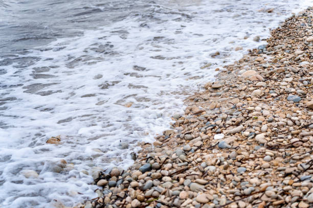 A serene coastline featuring smooth pebbles lining the shore, with gentle waves lapping at the water's edge under a clear blue sky on a sunny day.