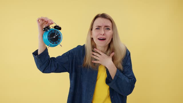 Emotional female in blue shirt pointing at clock with surprised facial expression on yellow studio background. Stressed woman touching head and complaining because of forgetting to watch time.