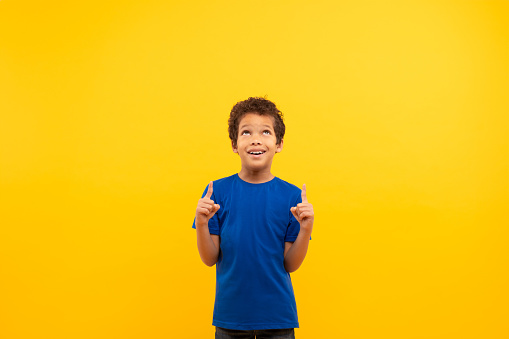 Curly-haired boy in blue t-shirt looking and pointing upwards at free space with look of curiosity set against bright yellow background, banner