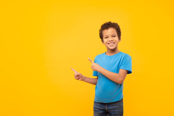 istock Joyful boy pointing to side at copy space, wearing blue t-shirt, yellow backdrop 1994798578