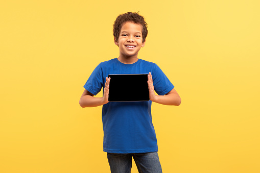 Cheerful young boy with curly hair, wearing blue t-shirt, proudly displays blank screen tablet with both hands, standing against vivid yellow background