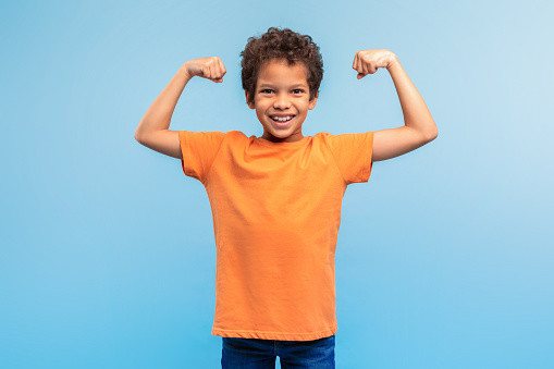 Young black boy in orange t-shirt showing off his muscles with big smile, exuding confidence and strength, against soft blue background